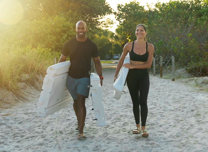 Man and woman walking on a sandy path with a stand up paddle board, surrounded by greenery.