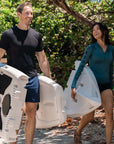 A man and woman carrying a light gray stand up paddle board outdoors with greenery in the background