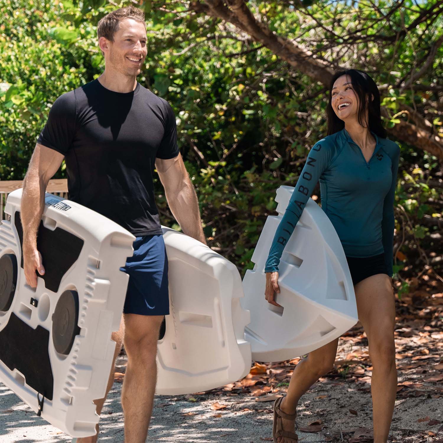 A man and woman carrying a light gray stand up paddle board outdoors with greenery in the background
